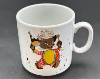 May include: White ceramic mug with a cartoon illustration of a brown and white animal wearing a red and white costume and playing a red and yellow drum. The animal is wearing a blue and yellow party hat. The background is white with blue and red stars.
