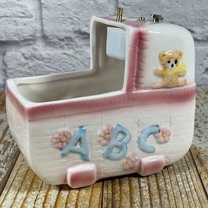 Può includere: Una scatola musicale in ceramica bianca a forma di culla per bambini con un design rosa e bianco. La scatola musicale ha un orsacchiotto sul lato e le lettere "ABC" sul davanti.