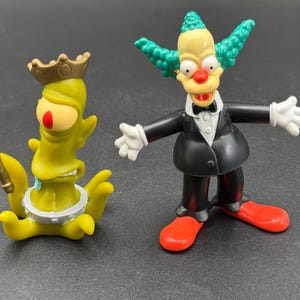 May include: Two cartoon character figurines. One is a green alien-like figure with a crown and scepter. The other is a clown with a black suit, red shoes, and green hair. Both figures are on a dark surface.