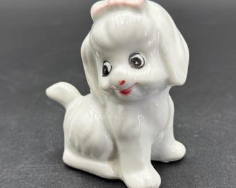 May include: A white ceramic figurine of a seated dog with a pink bow on its head. The dog has black eyes and a red mouth. The figurine is glossy and has a vintage aesthetic.