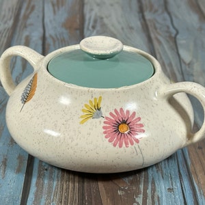 以下が含まれることがあります： 白地に緑がかった青色の蓋が付いた、花柄の白いセラミック製の砂糖入れ。砂糖入れには持ち手が2つあり、斑点模様の釉薬がかかっている。蓋の上には小さなつまみがある。