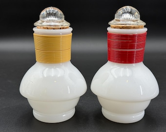 May include: Two white glass bottles with a bulbous shape. One has a yellow band, the other a red band, both around the neck. Each has a clear, textured glass stopper and a cork. The bottles are likely for spices or other small items.