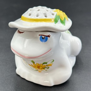 Può includere: Statuetta a forma di rana in ceramica bianca con cappello con finiture gialle e motivo a girasole. La rana ha occhi blu, labbra rosa e ciglia dipinte. L'oggetto è decorativo.