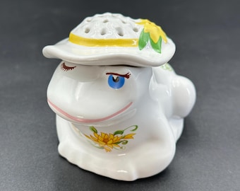May include: A white ceramic frog figurine wearing a hat with yellow trim and a sunflower design. The frog has blue eyes, pink lips, and painted eyelashes. The figurine is a decorative item.
