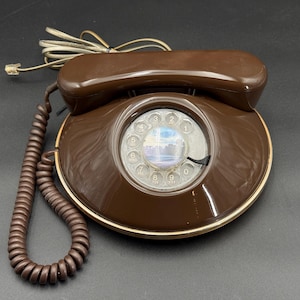 May include: A vintage brown rotary dial telephone with a coiled cord and a handset. The phone has a circular dial with numbers and a clear plastic cover. The base has a gold-coloured trim.