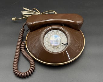 May include: A vintage brown rotary dial telephone with a coiled cord and a handset. The phone has a circular dial with numbers and a clear plastic cover. The base has a gold-colored trim.