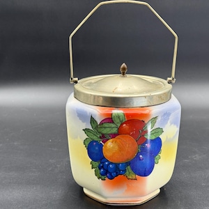May include: A vintage ceramic jar with a silver lid and handle. The jar is decorated with a colorful fruit design featuring red apples, orange and purple grapes, and green leaves. The background has a gradient of yellow, orange, and blue.