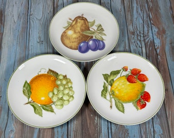 May include: Three white ceramic plates with green trim, each featuring a different fruit design. One plate has a pear, plums, and leaves, another has an orange, grapes, and leaves, and the last has a lemon, strawberries, and leaves.