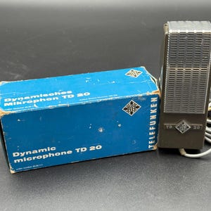 May include: Vintage Telefunken TD 20 dynamic microphone and its original blue box. The microphone is dark grey with a silver grill. The box has text in German and English, and the Telefunken logo.