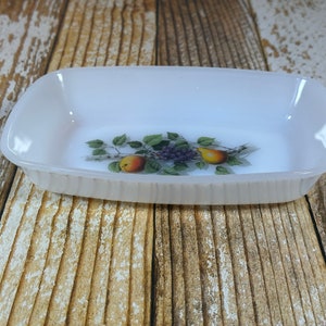 Puede incluir: Plato rectangular para hornear blanco con un diseño floral que presenta peras, uvas y hojas. El plato tiene un borde ligeramente ondulado.