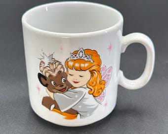 May include: White ceramic mug with a colorful illustration of a fairy with a crown and a furry animal friend. The fairy is wearing a white dress and has pink wings. The animal is brown and has a white star on its head.