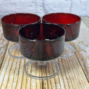 May include: Three red glass dessert cups with clear plastic bases. The cups are empty and sitting on a wooden surface.