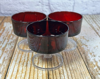 May include: Three red glass dessert cups with clear plastic bases. The cups are empty and sitting on a wooden surface.