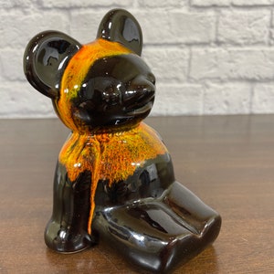 Puede incluir: Una figurita de cerámica de un oso con un esmalte negro y naranja. El oso está sentado con las piernas cruzadas y tiene una expresión amigable.
