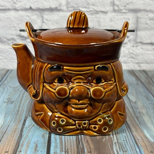 May include: A brown ceramic teapot shaped like a person with a moustache, glasses, and a bow tie. The teapot has a lid and a spout.