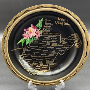 May include: A black decorative plate with a gold rim and a gold map of West Virginia. The plate features a floral design and the state's name. The plate is displayed on a clear stand.