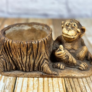 Puede incluir: Una maceta de cerámica marrón con forma de tocón de árbol con una figura de mono. La maceta tiene un aspecto texturizado similar a la madera. El mono está sentado, con una mano levantada, y tiene una expresión sonriente. La maceta está sobre una superficie de madera.