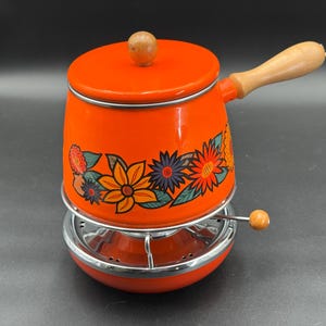 May include: An orange enamelware pot with a wooden handle and lid knob, decorated with floral patterns in yellow, orange, blue, and green. The pot sits on a chrome stand with a small wooden knob.