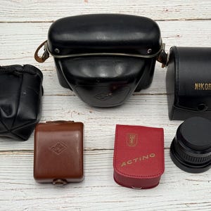 Peut inclure: Ensemble d'accessoires de photographie vintage. On distingue un grand étui en cuir noir, un petit étui noir avec "NIKORA" en lettres dorées et un étui rouge avec "ACTINO" imprimé. Sont également présents un objectif et une petite pochette noire.
