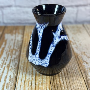 Puede incluir: Un jarrón pequeño de cerámica negra con un diseño abstracto blanco y azul. El jarrón tiene un cuerpo redondeado y una abertura estrecha.