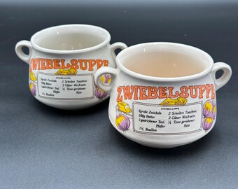 May include: Two white ceramic soup bowls with handles. The bowls have the words "ZWIEBELSUPPE" in orange lettering. The bowls also have a recipe for onion soup in German, with illustrations of onions and cheese.
