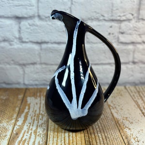 Puede incluir: Una jarra de cerámica negra con un asa curva y un diseño abstracto blanco. La jarra tiene un cuello delgado y una base redondeada. El diseño es una serie de líneas blancas que irradian desde la base.