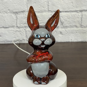 May include: A ceramic rabbit figurine painted in brown, red and grey. The rabbit has a grumpy expression with blue eyes and a white nose.