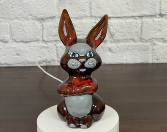 May include: A ceramic rabbit figurine with brown, red, and gray paint. The rabbit has a grumpy expression with blue eyes and a white nose.