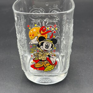 May include: A clear glass tumbler featuring a colourful graphic of Mickey Mouse dressed in safari gear with binoculars, surrounded by confetti and a red ribbon. The base of the glass is embossed with the year 2000, and the glass has embossed patterns.