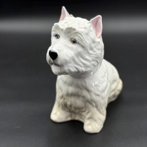Może przedstawiać: Ceramiczna figurka psa West Highland White Terrier. Pies jest biały z czarnym nosem i brązowymi oczami. Uszy mają różowe wnętrze. Figurka siedzi i ma puszysty wygląd.