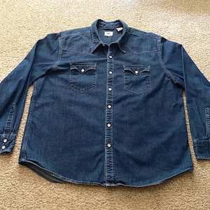 May include: A long-sleeved denim shirt in a medium blue wash. The shirt features two chest pockets with button closures, a button-down front, and buttoned cuffs. The collar is a classic point collar style.