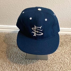 May include: A navy blue baseball cap with a white embroidered logo on the front. The cap has a curved brim and a button on top. The hat is displayed on a silver stand.