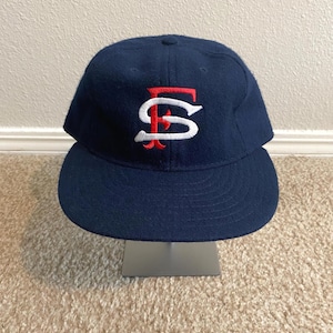 May include: A navy blue baseball cap with a curved brim. The cap features a white and red embroidered logo on the front. The hat is displayed on a silver stand against a neutral background.
