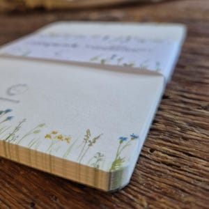 May include: A notepad with a floral design, featuring a cream-coloured cover adorned with illustrations of blue and yellow wildflowers and green grass. The pages are visible and have a light tan colour. The notepad rests on a wooden surface.