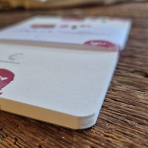 May include: Close-up of a white notebook with rounded corners. The pages are crisp and clean, with a slight texture. The notebook is partially open, revealing a glimpse of printed text and a red accent. The background is a wooden surface.