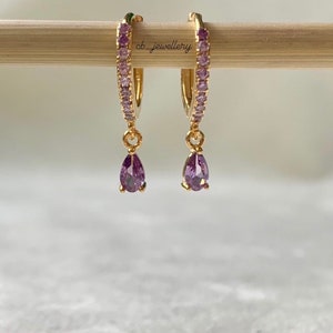 Könnte beinhalten: Goldene Creolen mit einer Reihe kleiner lila Steine um den Kreis und einem baumelnden birnenförmigen lila Stein.