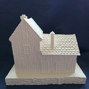May include: A white ceramic miniature house with detailed textured siding, roof tiles, and a chimney. The house features a front door, windows, and sits on a stone-like base. The overall design is a charming, rustic aesthetic.