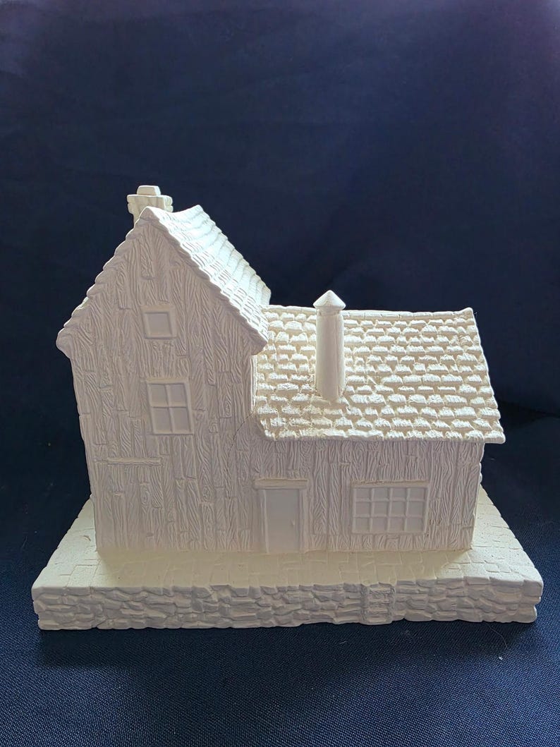 May include: A white ceramic miniature house with detailed textured siding, roof tiles, and a chimney. The house features a front door, windows, and sits on a stone-like base. The overall design is a charming, rustic aesthetic.