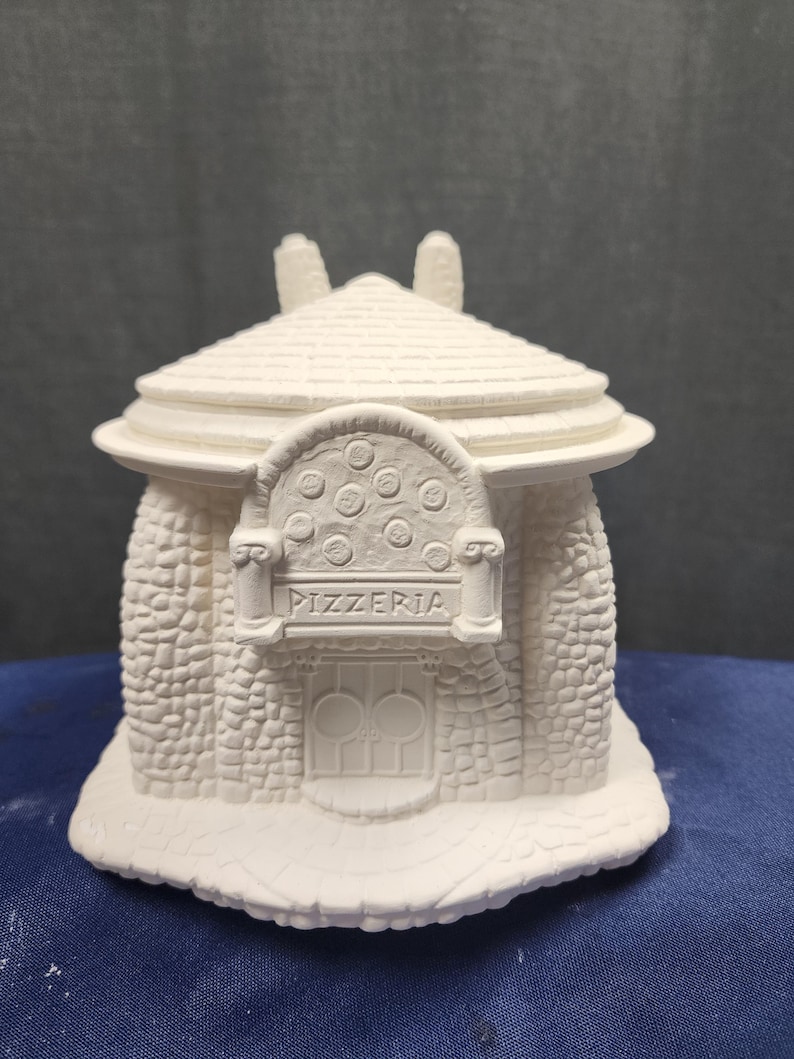 May include: White ceramic figurine of a small stone building with a rounded roof, a sign that reads "PIZZERIA", and a round window with a pizza design.