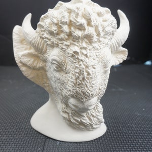 May include: White ceramic sculpture of a bison head with detailed fur texture. The sculpture is on a black surface.