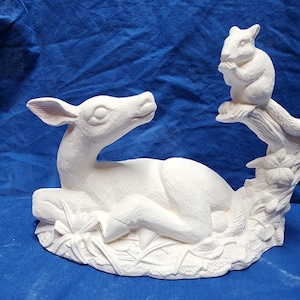 May include: A white ceramic figurine depicting a deer lying down, with a squirrel perched on a branch above. The deer is detailed with carved fur and rests on a bed of leaves. The squirrel holds a nut.