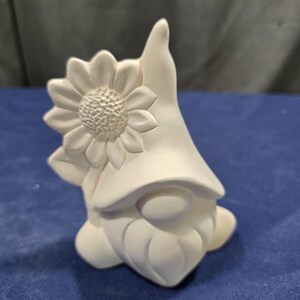 May include: A white ceramic gnome figurine featuring a large sunflower attached to the side of its tall, pointed hat. The gnome has a rounded nose and a textured beard, with a smooth, matte finish throughout the entire sculpted piece.