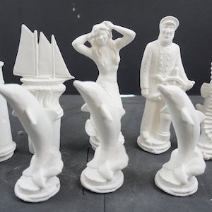 May include: A collection of unpainted ceramic nautical figurines, including a lighthouse, sailboat, mermaid, sailor, dolphin, and seahorse. The figurines are white and designed for painting or decorating.