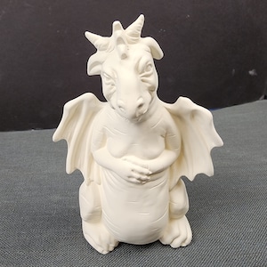 May include: White ceramic dragon figurine with wings, sitting with its arms crossed. The dragon has a pointed snout and a horn on its head.
