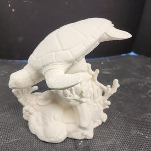 May include: A ceramic sculpture of a sea turtle, painted white, emerging from a coral reef base. The turtle's shell and flippers are detailed, and the coral has intricate textures. A decorative piece for coastal decor.