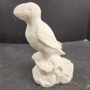 May include: White ceramic figurine of a puffin standing on a rock. The puffin is facing to the left and has a detailed beak and feathers.