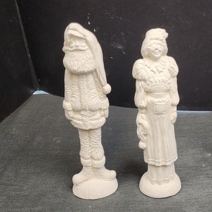 May include: Two white ceramic figurines, one depicting Santa Claus and the other a woman, both standing on a gray surface.