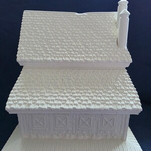 Puede incluir: Una figura de casa de cer&aacute;mica blanca con un tejado texturizado y dos chimeneas. La casa tiene una base rectangular y paneles decorativos en la parte delantera. El dise&ntilde;o es simple y r&uacute;stico.