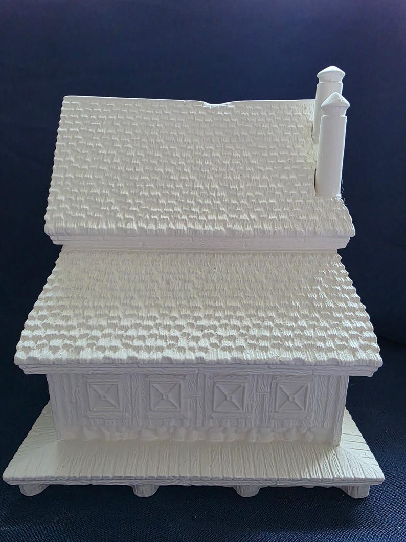 Puede incluir: Una figura de casa de cer&aacute;mica blanca con un tejado texturizado y dos chimeneas. La casa tiene una base rectangular y paneles decorativos en la parte delantera. El dise&ntilde;o es simple y r&uacute;stico.
