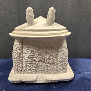May include: White ceramic house figurine with a stone-like texture and a small door. The house has two chimneys and a rounded roof.